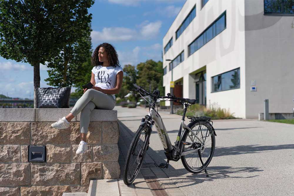 Eine Frau sitzt auf einer Mauer in der Stadt und neben ihr steht ein R Raymon e-Bike